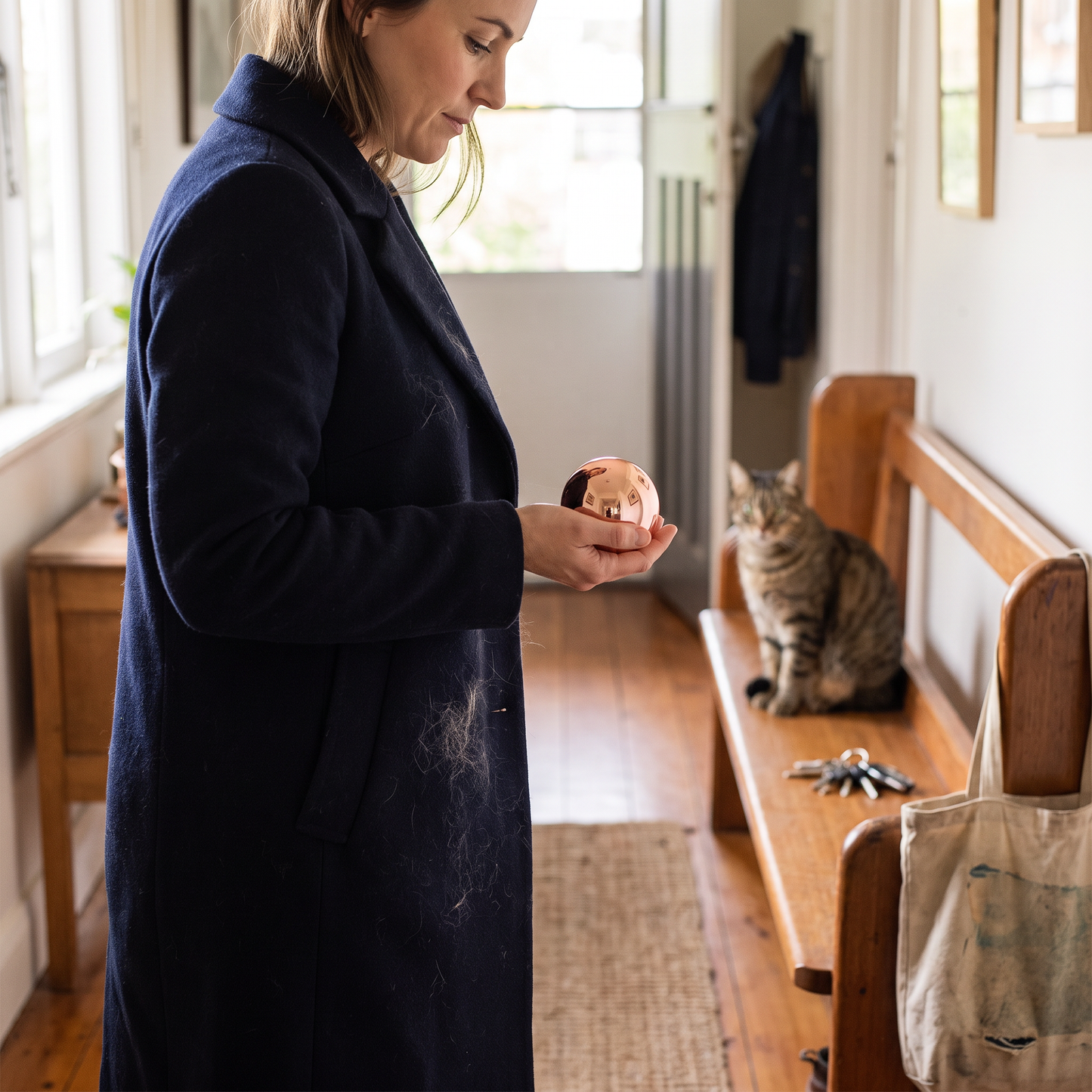 Woman in a navy coat by the front door using the Sidekick lint remover ball, cat on the hall bench